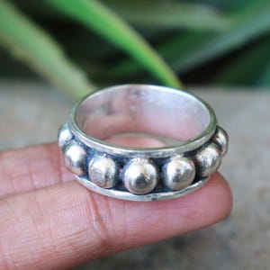 May include: A silver ring with a series of evenly spaced, raised, spherical accents around the band. The ring is shown close-up, with a blurred background of green foliage. The ring is a simple, yet elegant piece of jewelry.