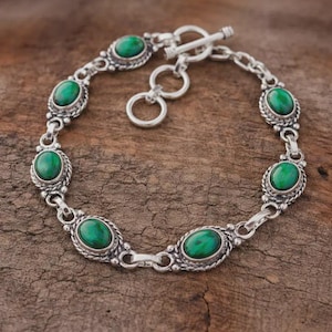 Könnte beinhalten: Ein silbernes Armband mit ovalen, leuchtend grünen Edelsteinen, die in detaillierten Fassungen gefasst sind. Das Armband hat einen Knebelverschluss und wird auf einer strukturierten, braunen Holzoberfläche präsentiert. Die Edelsteine sind gleichmäßig verteilt.