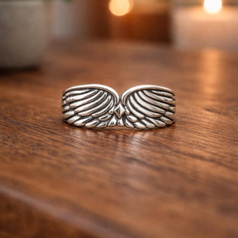 Peut inclure: Une bague argent&eacute;e orn&eacute;e d'ailes d&eacute;taill&eacute;es. La bague pr&eacute;sente deux ailes sym&eacute;triques avec des motifs de plumes d&eacute;taill&eacute;s, se rejoignant en un point central avec un accent en forme de diamant. La bague est pos&eacute;e sur une surface en bois chaleureuse.
