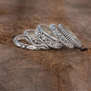 Könnte beinhalten: Nahaufnahme von fünf silbernen Ringen mit aufwendigen floralen und gemusterten Designs. Die Ringe sind auf einer Holzoberfläche gestapelt und zeigen die detaillierte Handwerkskunst. Jeder Ring hat ein einzigartiges Muster, einschließlich floraler, perlenbesetzter und gestreifter Texturen.