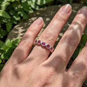 Könnte beinhalten: Ein Silberring mit einer Reihe von runden, in Zargenfassung gefassten, leuchtend pinkfarbenen Edelsteinen, die sich über die Vorderseite des Rings erstrecken. Das Metall hat eine glatte, polierte Oberfläche und die Steine sind in einer gleichmäßigen Reihe angeordnet.