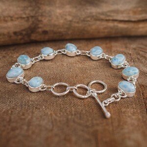 May include: A silver bracelet featuring oval, light blue gemstones. The gemstones are set in individual silver bezels and linked together with a silver chain. The bracelet has a toggle clasp for closure. The background is a brown wooden surface.