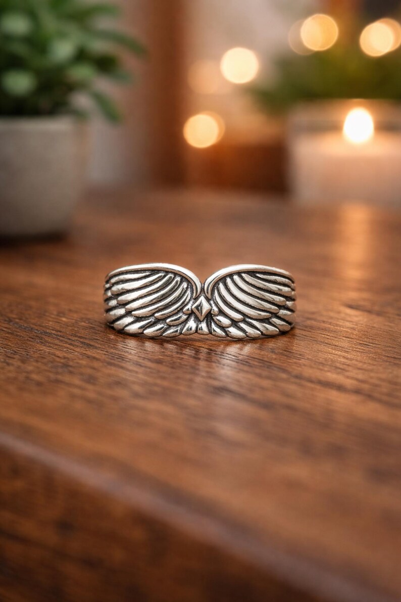 Peut inclure: Une bague argent&eacute;e avec un motif d'ailes. La bague pr&eacute;sente deux ailes d&eacute;taill&eacute;es qui se rejoignent au centre, avec un petit accent en forme de losange. La bague est pr&eacute;sent&eacute;e sur une surface en bois.