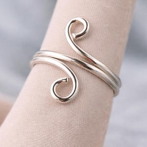 Könnte beinhalten: Ein silberfarbener Ring mit einem Doppelband-Design. Der Ring hat zwei wirbelnde, offen endende Spiralen, die sich um den Finger winden. Der Ring wird auf einem hellrosa Stoff mit einem weichen, verschwommenen Hintergrund präsentiert.