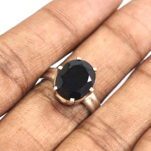 Könnte beinhalten: Ein Ring mit einem großen, oval geschliffenen schwarzen Edelstein, der in einer polierten, silberfarbenen Metallschiene gefasst ist. Der Stein wird von sechs kleinen Krappen gehalten, und die Schiene hat eine glatte, schlichte Oberfläche.