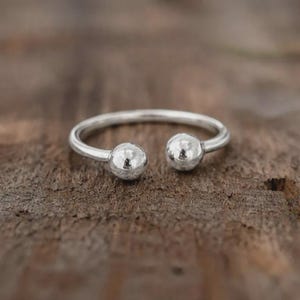 Könnte beinhalten: Ein silberner offener Ring mit zwei kugelförmigen Enden. Der Ring liegt auf einer dunklen, strukturierten Holzoberfläche, die das polierte Metall und das minimalistische Design des Rings hervorhebt. Das Design des Rings ist modern.
