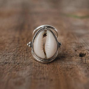Könnte beinhalten: Ein silberner Ring mit einer großen, cremefarbenen Kaurischnecke. Die Schnecke ist in einem detaillierten, verzierten Silberband mit einem gedrehten Seildesign eingefasst. Der Ring hat eine Vintage- oder Boho-Ästhetik.