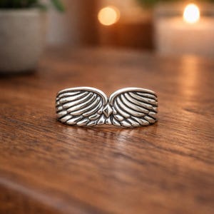 Peut inclure: Une bague argent&eacute;e avec un motif d'ailes. La bague pr&eacute;sente deux ailes d&eacute;taill&eacute;es qui se rejoignent au centre, avec un petit accent en forme de losange. La bague est pr&eacute;sent&eacute;e sur une surface en bois.