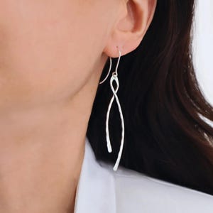 Könnte beinhalten: Silberfarbene, längliche Ohrhänger mit gehämmerten Strukturen. Die Ohrringe haben ein geschwungenes, fließendes Design mit Hakenverschluss. Ein schlichtes, elegantes Accessoire.
