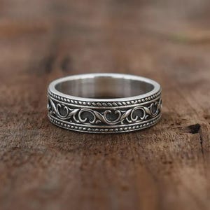 Könnte beinhalten: Ein silberner Ring mit einem verzierten, sich wiederholenden Rankenmuster. Der Ring weist ein detailliertes Muster aus wirbelnden Linien und Kurven auf, das von einem Perlenrand begrenzt wird. Der Ring ist vor einem hölzernen Hintergrund platziert.