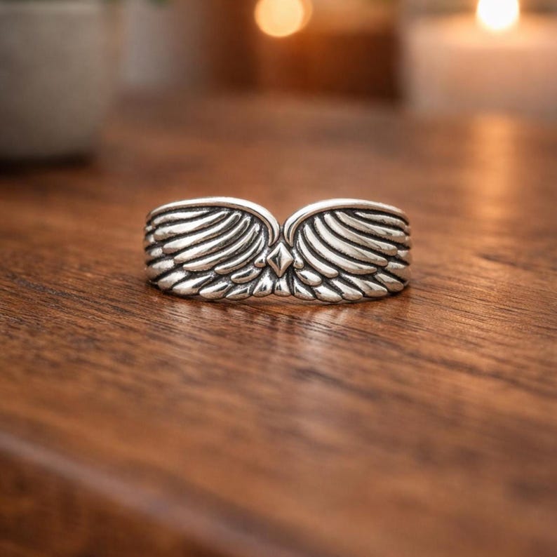 Peut inclure: Une bague argent&eacute;e avec un motif d'ailes. La bague pr&eacute;sente deux ailes d&eacute;taill&eacute;es qui se rejoignent au centre, avec un petit accent en forme de diamant. La bague est pos&eacute;e sur une surface en bois.