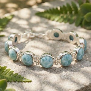 Könnte beinhalten: Ein Silberarmband mit einer Reihe runder, polierter hellblauer Larimar-Edelsteine, die in glatte Metallfassungen eingefasst sind. Das Armband besteht aus abwechselnden kreisförmigen Steinfassungen und kleinen silbernen Verbindungsgliedern.