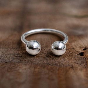 May include: A silver-colored open ring with two spherical ends. The ring is made of a shiny metal and sits on a wooden surface. The design is simple and modern, suitable for everyday wear.