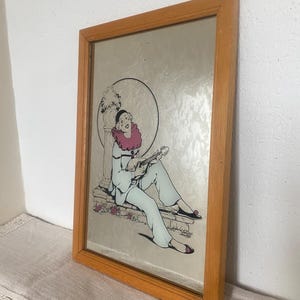 Peut inclure: Miroir encadré avec une illustration d'une personne jouant d'un instrument à cordes. La personne porte une tenue bleu clair, un col à volants rose et des chaussures noires. L'œuvre est encadrée dans un cadre en bois.