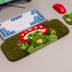 Peut inclure: Un repose-poignet rectangulaire touffeté pour clavier et un repose-poignet assorti en forme de grenouille pour souris. Les deux présentent une surface verte texturée avec un motif touffeté de grenouille sous un champignon rouge et blanc.
