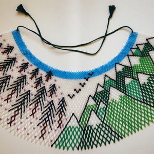 May include: A beaded bib necklace with a landscape design. The necklace has a white base with black trees and green mountains. A blue beaded band forms the neckline, and black cords with tassels are attached.