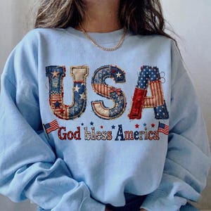 Puede incluir: Sudadera azul claro con la palabra "USA" en un diseño de patchwork de estrellas y rayas rojas, blancas y azules. La frase "God bless America" está bordada debajo, con pequeñas banderas americanas.