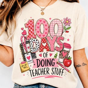 May include: A cream-coloured t-shirt featuring a graphic with the text 100 DAYS OF DOING TEACHER STUFF. The design includes pink and red patterns, a notebook, a teacher ID badge, a coffee cup, a red apple, a crayon, and a yellow sticky note.