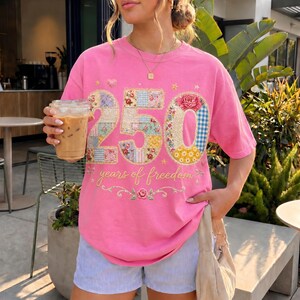 May include: A pink t-shirt with the number "250" in a patchwork design, with the words "years of freedom" below. The shirt is worn with blue and white striped shorts. A person is holding a clear plastic cup with a brown beverage.