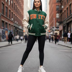 May include: A green and cream varsity jacket with the word "UNITY" in orange lettering. The jacket has white stripes on the cuffs and hem. The person is wearing black leggings and white trainers.