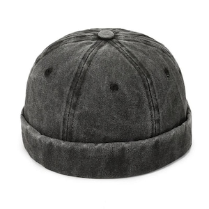 May include: A dark gray denim cap with a rolled brim. The cap has a rounded crown with a button on top and stitched panels. The fabric has a faded, worn appearance, suggesting a vintage or distressed style. The cap is a classic, casual accessory.