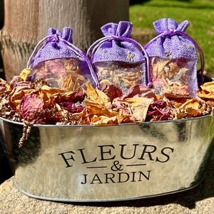 May include: Three small purple bags filled with dried flower petals are displayed in a silver metal container with the words "FLEURS & JARDIN" printed on the side.