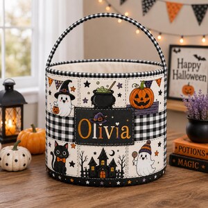 May include: A cylindrical fabric basket with a black and white gingham handle and trim. The exterior features a grid pattern with illustrations of a ghost, a pumpkin, a black cat, a haunted house, and a cauldron. The name Olivia is printed in orange.