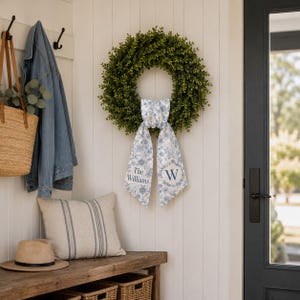 May include: A round green boxwood wreath featuring a hanging fabric ribbon with a blue and white floral pattern. The ribbon displays the text The Williams and the letter W in a dark blue font, centered on the wreath against a white wall.