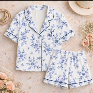 May include: White pajama set with a blue floral pattern, featuring a button-down shirt with short sleeves and matching shorts. The set has blue trim and an embroidered "M" on the shirt. A comfortable sleepwear option.