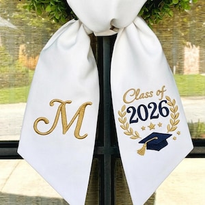 May include: White graduation stole featuring a gold embroidered initial "M" and the text "Class of 2026" with a graduation cap and laurel wreath design. The stole is displayed in front of a window.