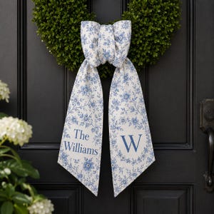 Op de afbeelding: Een decoratieve stoffen strik met lange hangende uiteinden voorzien van een blauw-wit bloemenpatroon. Het linker uiteinde toont de tekst The Williams, terwijl het rechter uiteinde een grote letter W in een cirkelvormige bloemenkrans bevat.