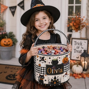 May include: A cylindrical fabric Halloween candy bucket with a black and white buffalo check pattern on the rim and handle. The exterior features colourful illustrations of ghosts, a pumpkin, a black cat, and a haunted house, with the name Olivia.