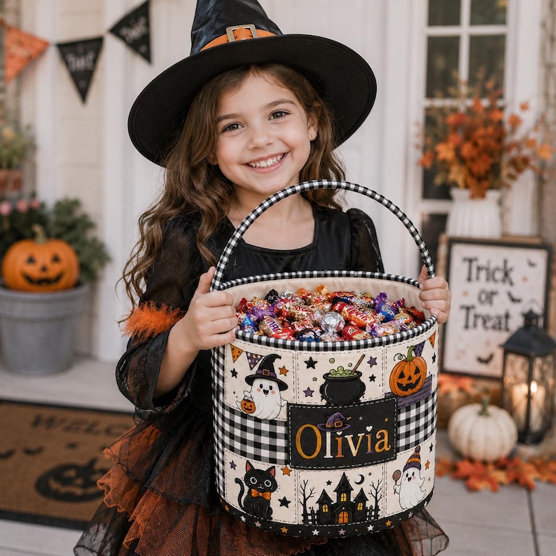 May include: A cylindrical fabric Halloween candy bucket with a black and white buffalo check pattern on the rim and handle. The exterior features colourful illustrations of ghosts, a pumpkin, a black cat, and a haunted house, with the name Olivia.