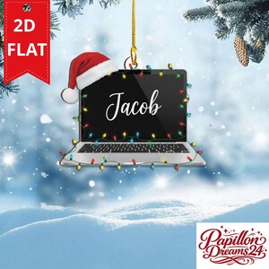 May include: A flat, two-dimensional hanging ornament shaped like a silver laptop computer. The screen displays the name Jacob in white script. A red and white Santa hat sits on the top left corner, and a string of colourful Christmas lights surrounds it.