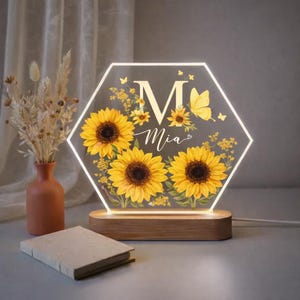 Puede incluir: Una lámpara LED personalizada con forma hexagonal. La lámpara presenta un diseño de girasoles con la letra "M" y el nombre "Mia". La base es de madera. La lámpara es un artículo decorativo.
