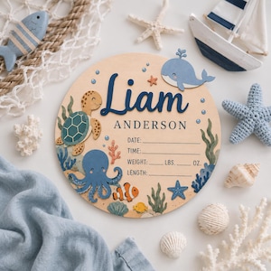 May include: A circular wooden birth announcement sign featuring the name Liam Anderson in dark blue script. The design includes illustrations of a sea turtle, octopus, whale, and clownfish, with blank lines for date, time, weight in lbs and oz, and length.