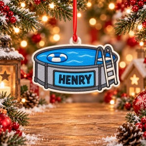 Könnte beinhalten: Ein flacher, ovaler Weihnachtsbaumschmuck mit einer Grafik eines grauen Aufstellpools mit blauem Wasser, einem weiß-blauen Rettungsring und einer Leiter. Der Name HENRY ist in fetten, schwarzen Großbuchstaben auf der Poolwand abgedruckt.