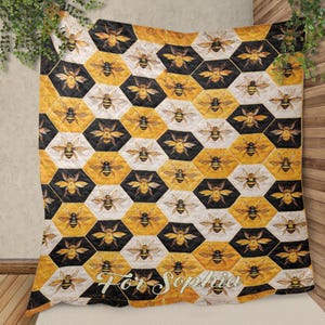 May include: A quilted blanket with a repeating pattern of bees inside of honeycomb-shaped cells. The cells alternate between black, white, and yellow. The blanket has a decorative border and the text "For Sophia" in a script font.
