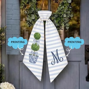 May include: A door hanging with a blue and white striped design. It features a topiary in a blue and white pot, decorated with bows, and a large letter "M". The hanging is suspended from a chain.