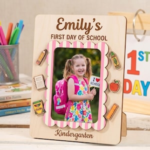 Peut inclure: Un cadre photo en bois clair portant le texte Emily's FIRST DAY OF SCHOOL et Kindergarten. Le cadre présente une bordure festonnée à rayures roses et blanches, ornée d'icônes en bois représentant un crayon, un cahier, un crayon de cire, une pomme et une règle.