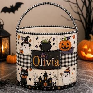 May include: A cylindrical Halloween bucket with a black and white buffalo check handle and trim. The exterior features a cream-coloured fabric panel with illustrations of ghosts, a pumpkin, a black cat, a cauldron, and a house. The name Olivia is printed in orange on a central black rectangular patch.