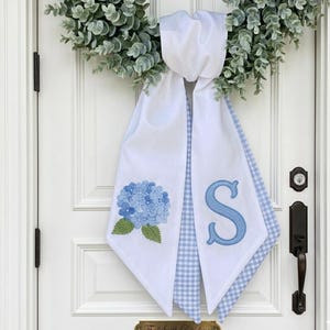 Puede incluir: Bufanda blanca para puerta con bordado de hortensia azul y letra "S" azul. La bufanda tiene un estampado de cuadros vichy azul claro en los bordes. Una corona verde cuelga encima. La puerta tiene una placa de latón.