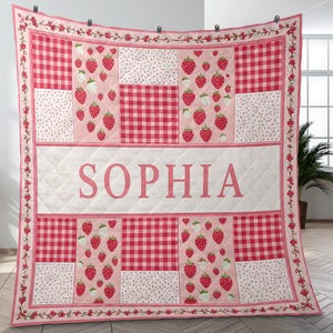 May include: A personalised quilt with a patchwork design. It features red gingham, strawberry patterns, and floral accents. The name "SOPHIA" is displayed in the centre. The quilt has a pink and white colour scheme.