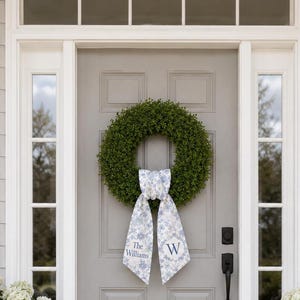 May include: A circular green boxwood wreath featuring a decorative fabric ribbon tied at the bottom. The ribbon has a light blue and white floral pattern with the text The Williams on the left tail and the letter W on the right tail.