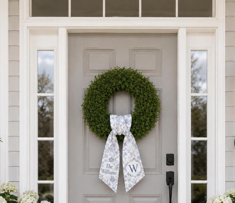 May include: A circular green boxwood wreath featuring a decorative fabric ribbon tied at the bottom. The ribbon has a light blue and white floral pattern with the text The Williams on the left tail and the letter W on the right tail.