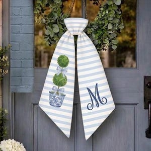May include: A decorative door hanging with blue and white stripes. One side features a topiary design in a blue and white planter, and the other side has a cursive "M" in navy blue. The hanging is suspended by a chain.