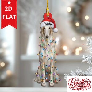 May include: A flat 2D hanging ornament featuring a white Borzoi dog wearing a red and white Santa hat with the name Teddy printed on the brim. The dog is wrapped in a string of colorful Christmas lights, including blue, red, and yellow bulbs.
