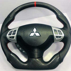May include: Black and carbon fiber steering wheel with red stitching and a red stripe at the top. The center features the Mitsubishi logo and silver control buttons. The wheel has a perforated leather grip.