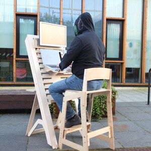 May include: A light-colored wooden standing desk with a matching chair. The desk has a monitor stand, a laptop surface, and a lower shelf. A person is using the desk outdoors, wearing a black hoodie and jeans.