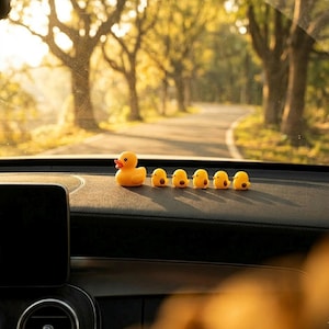 Puede incluir: Un pato de goma amarillo y cinco patitos más pequeños están colocados en el salpicadero de un coche. Los patos miran hacia delante, con una carretera y árboles visibles a través del parabrisas. La escena está bañada por la luz del sol.